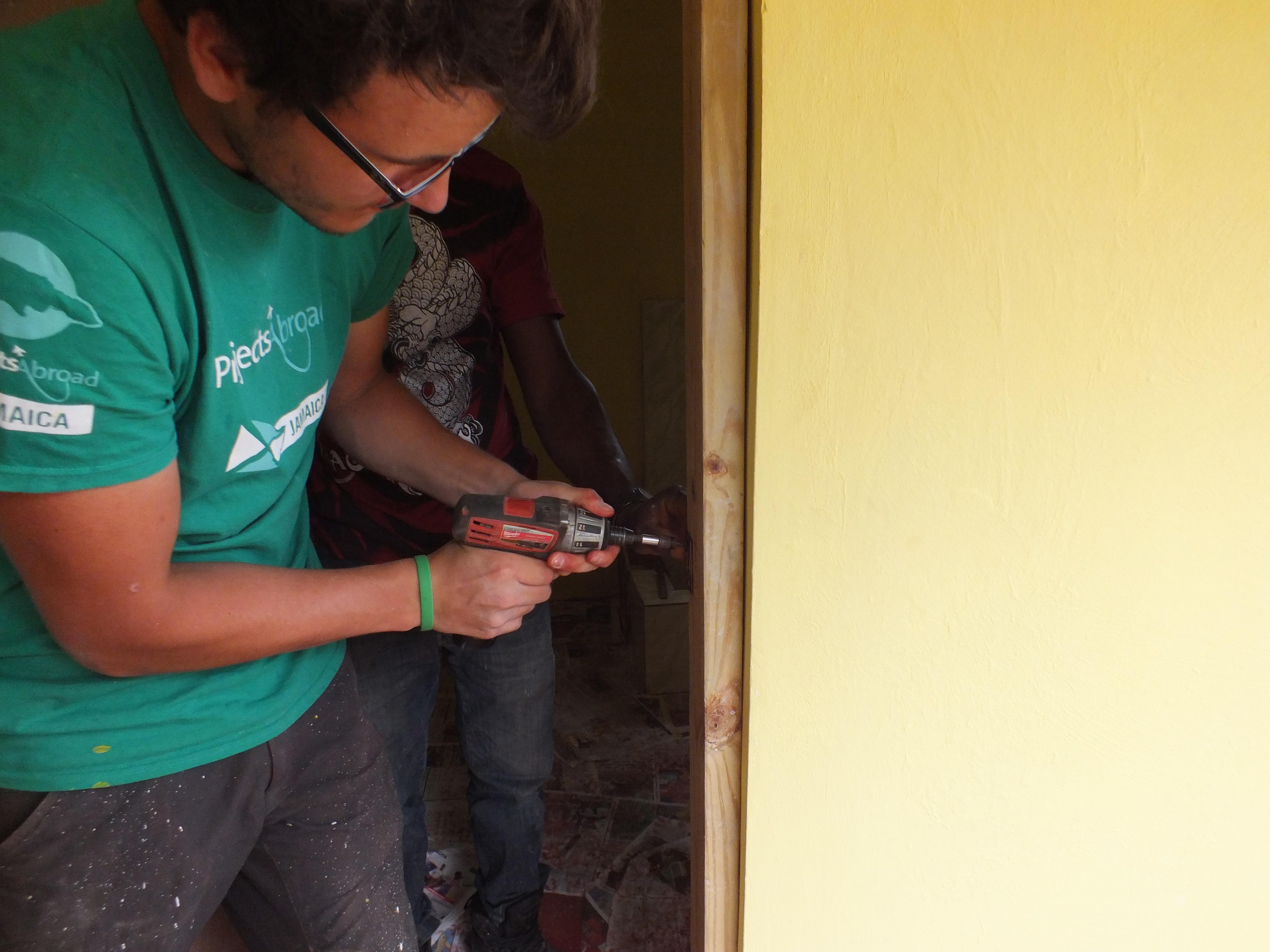 A Projects Abroad volunteer working with a group attaches a door to a door frame as part of building volunteer work in Jamaica. A volunteer helps with building a bathroom as part of construction volunteering in Jamaica for a group.